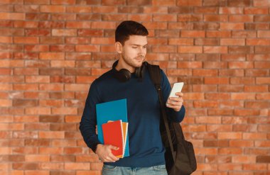 Students Lifestyle. Millennial Guy Using Smartphone Standing Over Brick Wall Background Indoor