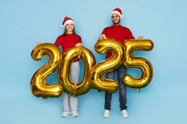 Kırmızı süveter ve Noel Baba şapkalı bir erkek ve kadın 2025 yılını gösteren büyük altın balonları neşeli bir şekilde tutuyorlar..