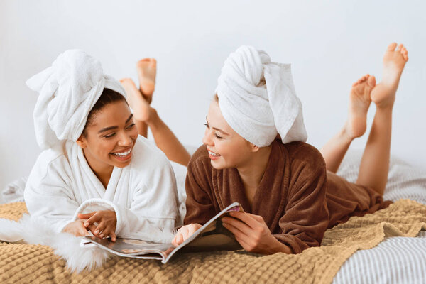 Young beautiful multiracial women in bathrobes and towels on head happily talking and reading magazines together while laying on bed at home