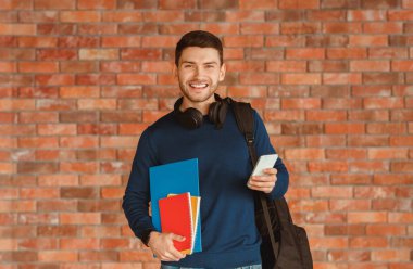 Modern Student. Happy Guy With Backpack Holding Smartphone And Notebooks Smiling To Camera Standing Over Brick Wall Background Indoor clipart