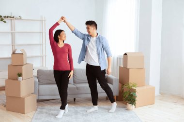 Taşınma gününü kutluyoruz. Mutlu Asyalı genç kiracılar yeni evlerinde paketsiz kutuların arasında dans ediyorlar.
