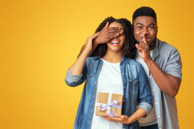 Sürpriz hediye. Kız arkadaşının gözlerini kapayan ve doğum günü ya da Noel hediyesi verirken sessizlik işareti gösteren, sarı arka plan.