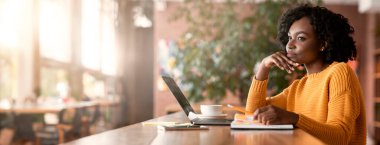 İnternette yeni bir iş arayan düşünceli afro genç kadın, kafede dizüstü bilgisayar kullanıyor, notlar alıyor, yan görüş alıyor, boşlukları kopyalıyor.