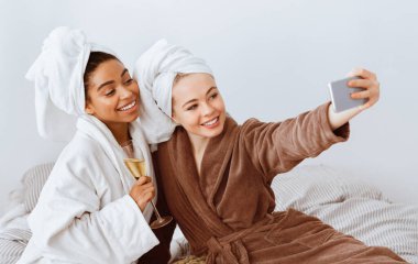 Multiethnic cheerful ladies taking selfie in bathrobes, leisure at home concept