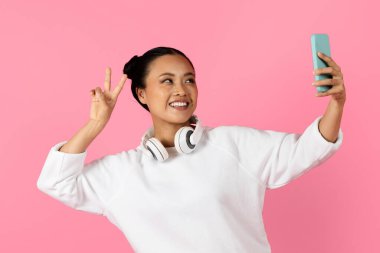 Asyalı kadın telefonuyla selfie çekerken ışıl ışıl gülümsüyor. Boynuna beyaz kulaklık takıyor, yumuşak pembe bir arka plana şakayla poz veriyor..