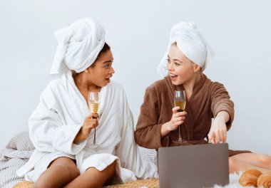 Evde spa yapan, şampanya içen ve dizüstü bilgisayar kullanan rahat kız arkadaşlar, internette güzellik teklifleri.