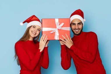 Kırmızı giyinmiş neşeli bir çift ve Noel Baba şapkaları güzel paketlenmiş bir Noel hediyesini tutarken gülümsüyor. Neşeli atmosfer tatil sezonunun ruhunu yansıtıyor..