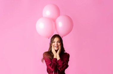 Neşeli ruh hali. Üç pembe balon tutan neşeli genç bayan heyecanla yanaklarına dokunuyor, renkli arka planda poz veriyor.