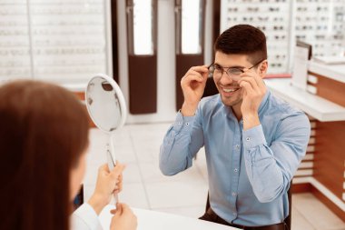Göz Giysisi Konsepti Seçimi. Kadın optometrist yakışıklı bir adamın ayna tutarak gözlük seçmesine yardım ediyor.
