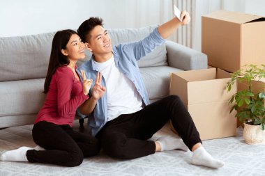 Yeni Ev, Teknoloji Konsepti. Mutlu Asyalı çift akıllı telefonlu selfie çekiyor. Karton kutuların arasında oturuyorlar.