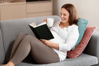 Genç hamile bir kadın rahatça koltukta oturur, çay içerken kitap okumaya dalmıştır. Modern bir yaşam alanında rahat ve mutlu görünüyor..