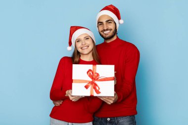 Kırmızı kazaklar ve Noel Baba şapkaları giyen neşeli bir çift ellerinde güzel bir hediye, bir kurdeleyle süslenmiş, Noel ve beraberliğin neşesini somutlaştırıyorlar..