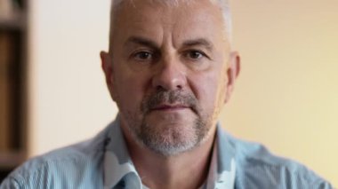 Olgun bir adam evde sakince oturur, düşünceli bir şekilde yüzüne vurgu yaparak yüzüne bakar..