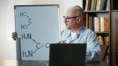 Profesör, masa başında otururken beyaz tahtadaki moleküler yapıları göstererek organik kimya öğretiyor..