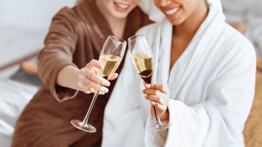 İki etnik çeşitlilikte spa kız arkadaşı şampanya kadehleriyle tezahürat yapıyor, bornoz giyiyorlar.