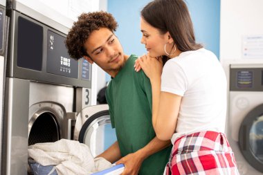 A young couple shares a moment at a laundromat, with one partner loading clothes into a washer while the other looks over affectionately, creating a warm and familiar atmosphere. clipart