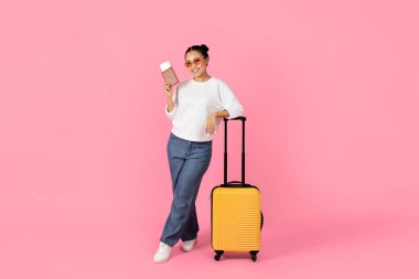 Neşeli genç Asyalı bir kadın elinde akıllı bir telefonla parlak sarı bir bavulun yanında duruyor. Günlük kıyafetler ve güneş gözlüğü takıyor. Pembe bir arka plana rahat bir seyahat havası yayıyor..