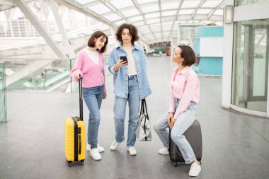 Üç kadın bir havaalanı terminalinde toplanırken, ikisi bavullarının yanında duruyor. Sohbete dalmış gibi görünüyorlar ve bir akıllı telefondan detayları kontrol ediyorlar..
