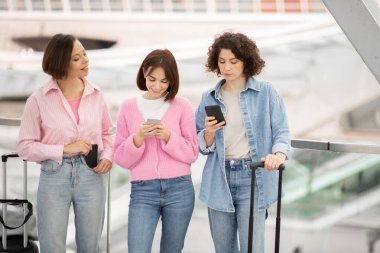 Çağdaş bir havaalanı ortamında üç kadın yan yana duruyor, her biri cep telefonlarına dalmış. Biri bavulu tutarken, diğerleri de cihazlarını kontrol edip, seyahat aktivitelerini gösteriyor..