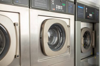 Three stainless steel washing machines are lined up in a laundromat, showcasing their circular glass doors. The bright interior highlights the machines in use during the day. clipart