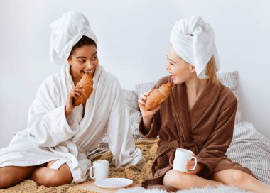Neşeli, çoklu etnik spa kız arkadaşları yatakta birlikte kahvaltı yapıyor, taze kruvasan yiyor, kahve içiyor.