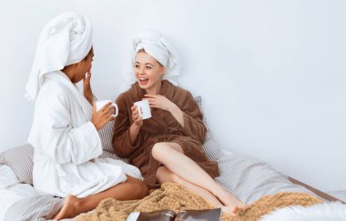 Yatakta oturup çay içen, sohbet eden, boş alan arayan, bornozlu neşeli çok ırklı kız arkadaşlar.