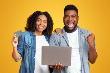 Çevrimiçi kolay ödemeler. Başarılı Afrikalı çiftin portresi sarı arka planda dizüstü bilgisayar ve kredi kartıyla başarıyı kutluyor.