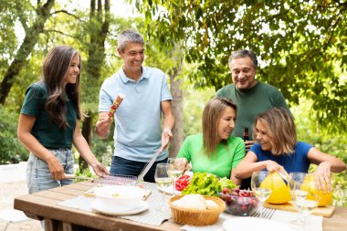 Neşeli bir arkadaş grubu bir masanın etrafında toplanıyor, ızgara yaparken yemek ve kahkaha paylaşıyorlar. Atmosfer canlı, içecekler ve çeşitli yemekler var..