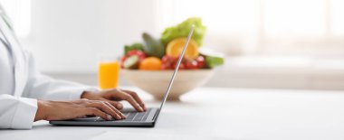 African woman nutritionist doctor working on laptop at office, bowl with fruits and glass of juice on table, cropped, copy space, side view clipart