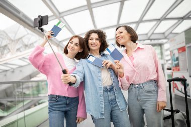 Üç arkadaş havaalanında keyifli bir anın tadını çıkarıyorlar. Biniş kartlarını tutarken selfie çekiyorlar. Birlikte yapacakları yolculuk için heyecanlılar..