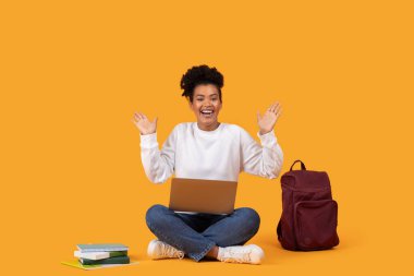 Neşeli genç bir kadın dizüstü bilgisayarı, kitapları ve sırt çantasıyla yerde bağdaş kurup parlak turuncu bir fon karşısında başarılarını kutluyor..