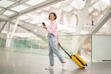 Pembe bluzlu ve açık renk kotlu genç bir kadın havaalanı terminalinden geçerken telefonunda güncelleme ya da bilgi kontrolü yaparken neşeli sarı bir bavul taşıyor..