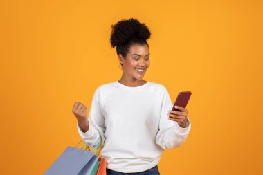 Genç bir kadın parlak turuncu bir arka plana karşı duruyor, bir elinde renkli alışveriş torbaları, diğer elinde bir telefon, heyecanla mesajlarını kontrol ederken gülümsüyor..
