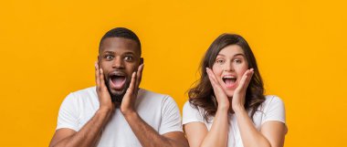 Harika satışlar. Şok olmuş ırklar arası bir çift kameraya bakıyor ve yanaklarına hayretle dokunuyor, sarı arka planda birlikte poz veriyorlar.