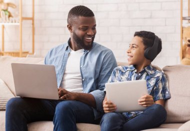 Aile Aygıtları. Neşeli Kara Baba ve Oğlu Dijital tablet ve dizüstü bilgisayarlarını kullanarak koltukta dinleniyorlar, interneti geziyorlar, boş yerleri geziyorlar.