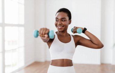 Ağırlıklarla Aile Eğitimi. Pozitif siyahi kadın dambıllarla egzersiz yapıyor, vücudunu evde güçlendiriyor. Diş teli takmış, kaslarını çalıştıran, sağlıklı kalan gülümseyen genç bir kadın.