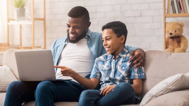 Genç siyah baba ve küçük oğlu evde dizüstü bilgisayar kullanıyor, bilgisayar ekranına bakıyor, online alışveriş yapıyorlar.