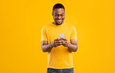 Gülümseyen Siyah Adam Smartphone Tarama İnternet ve Sosyal Medya Haber Kaynağı Sarı Stüdyo Arkaplanı 'nın üzerinde duruyor. Millennial Guy Yeni Mobil Uygulama Kullanıyor. İnsanlar ve Gadget