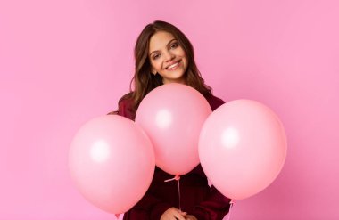 Pozitif genç kadın üç pembe balonla poz veriyor, renkli arka planda duruyor ve kameraya gülümsüyor, boş alan.