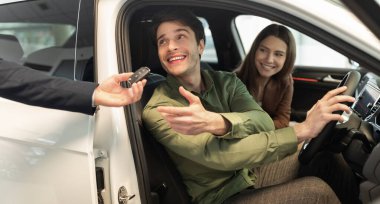 Happy millennial couple taking car key from auto salesman, sitting inside modern automobile at dealership, panorama. Cheery young family buying new vehicle at modern showroom