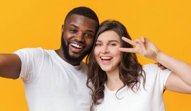 Uluslararası arkadaşlar. Neşeli siyah adam ve beyaz kız selfie çekiyorlar ve barış işareti gösteriyorlar, sarı arka planda eğleniyorlar.