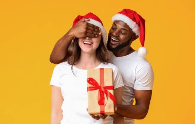 Sürpriz Noel Hediyesi. Noel Baba şapkalı Afrikalı Amerikalı adam, beyaz kız arkadaşının gözlerini kapayıp Noel hediyesi veriyor. Sarı arka plan.