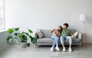 Spending weekend together. Happy millennial Asian couple resting on couch at home, empty space. Cheerful young spouses relaxing and enjoying each others company in living room