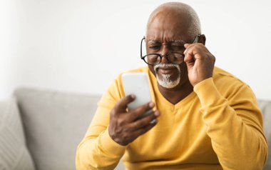 Zavallı görme duyusu. Evde koltukta oturan gözlüklü, akıllı telefon kullanan kıdemli siyah adam. Kötü Görüş, Göz Sağlığı ve Sağlık Sorunları Konsepti. Seçici Odaklanma