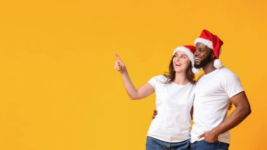 Noel teklifi. Noel Baba şapkası takan mutlu bir çift sarı arka plandaki fotokopi alanına bakıyor ve işaret ediyor.