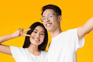 Neşeli Asyalı genç çift selfie çekiyor, kız barış işareti gösteriyor, sarı stüdyo geçmişi var.