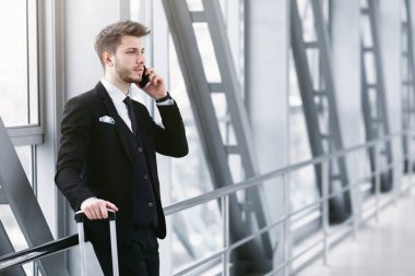 Ciddi Sakallı Girişimci Telefonla Konuşuyor ve Havaalanında Ayrılışını Bekliyor, Bavulu Tutuyor