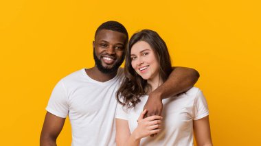 Happy multiracial couple hugging and posing over yellow background, affectionate black man embracing his girlfriend, empty space
