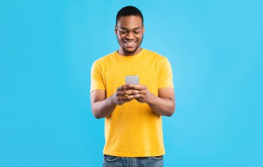 Mavi Stüdyo Arkaplanının Üzerinde Duran Cep Telefonu Mesajı Kullanarak Afro-Amerikalı Mutlu Erkek. Neşeli siyahi adam internette geziniyor. Akıllı telefonunuz için yeni bir uygulama. İnsanlar ve Gadget