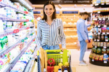 Gülümseyen Neşeli Genç Kadın markette raflarda alışveriş arabasıyla geziyor. Marketten alışveriş yapan, mandıralı dondurucuya bakan mutlu kadın müşteriler.